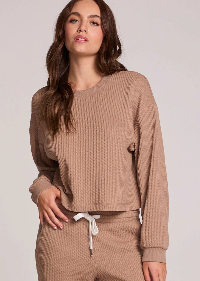 A woman modeling a matching ribbed lounge set in a warm taupe-brown color, featuring a cropped long-sleeve top and coordinating pants with a white drawstring, against a plain light gray studio background.